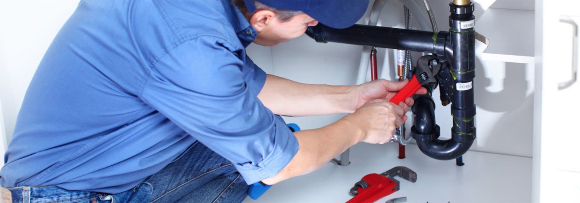 A plumber repairs black plastic pipes under a sink using a red pipe wrench.