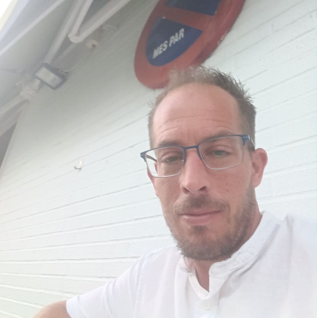 Man with glasses and a beard wearing a white shirt against a white brick wall.