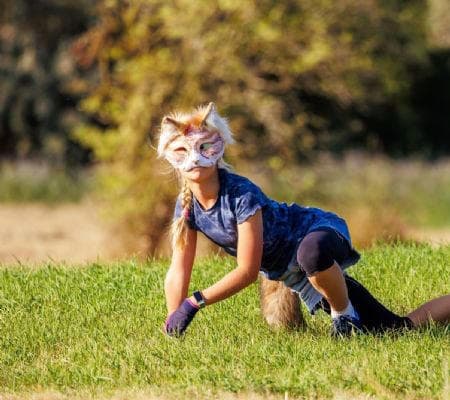 Girl wearing a fox mask and tail crouches on all fours in a sunny field.