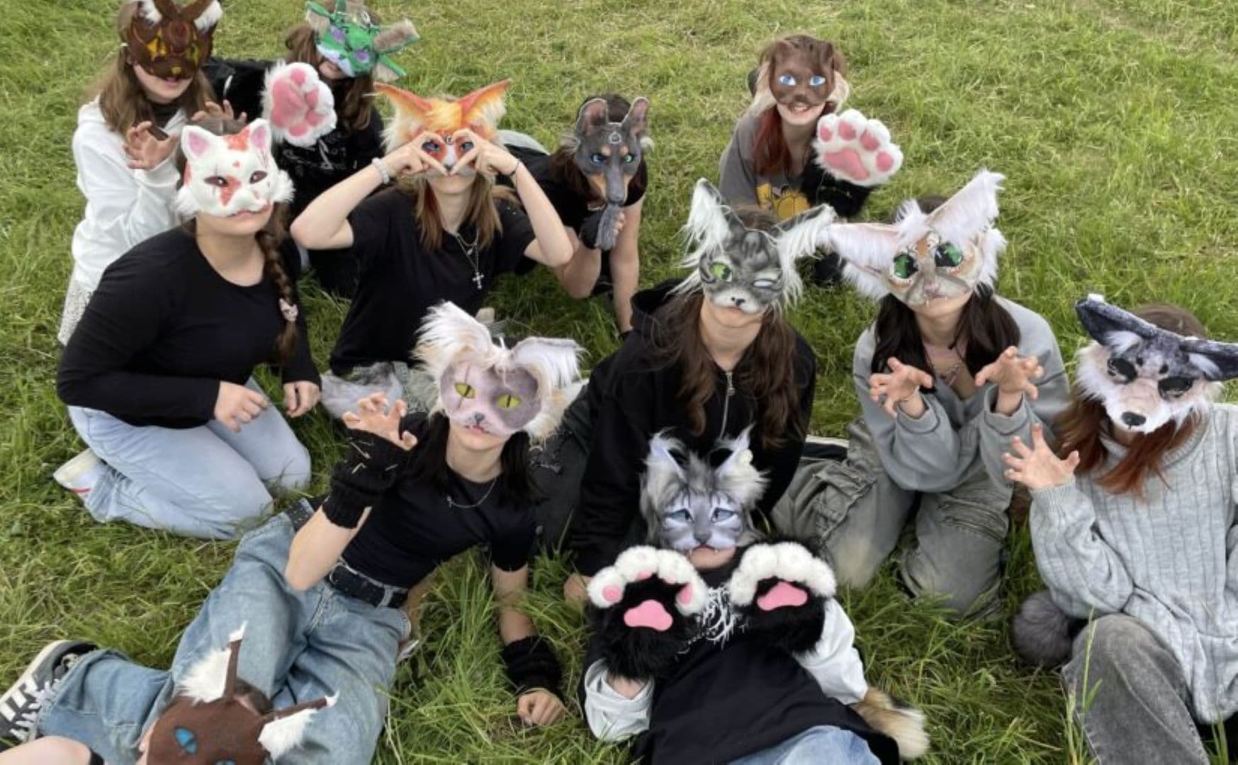 Teenagers wearing stylized animal masks and paw gloves pose in a green grassy field.