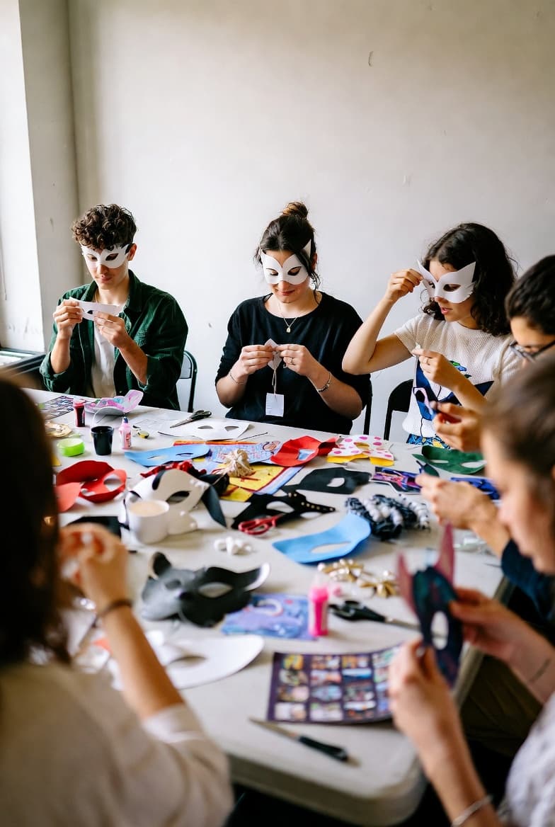 People wearing white masks sit at a table making more masks with various craft supplies.