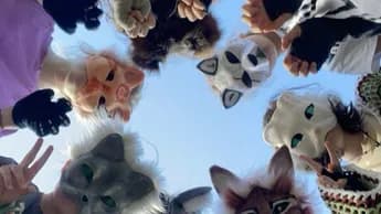 A circle of people wearing animal masks look down into the camera against the sky.