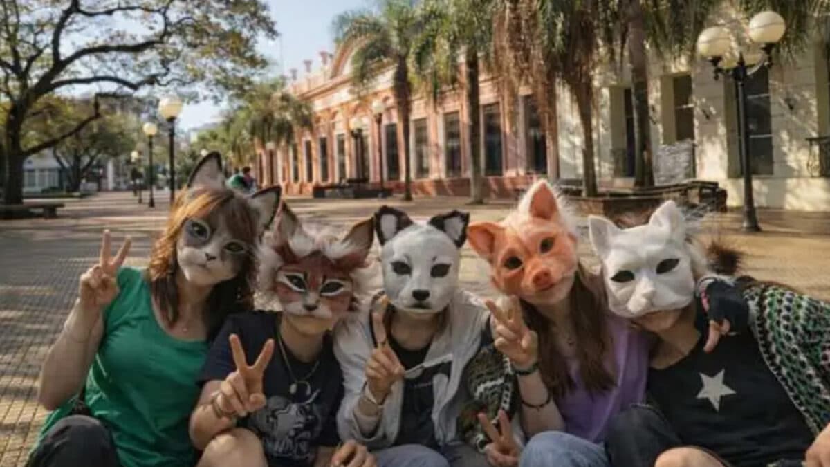 Five people wearing realistic animal masks sit in a sunny plaza, making peace signs.