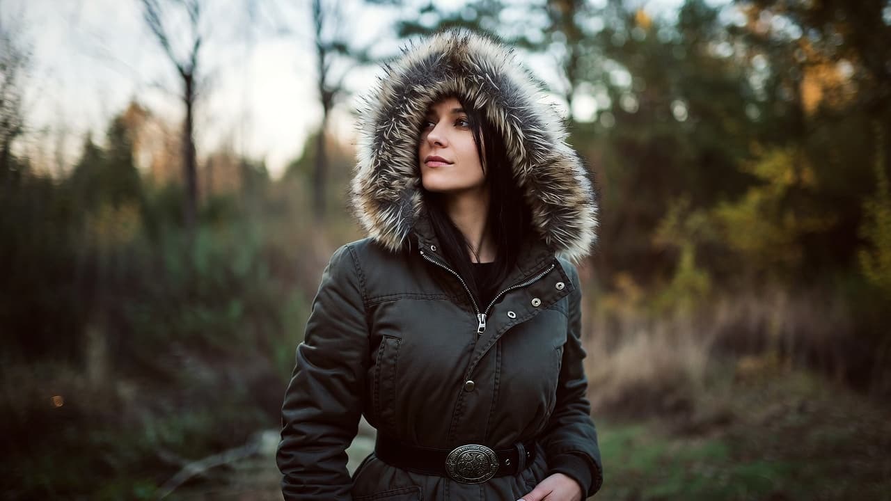 Woman wearing a green fur-trimmed parka and ornate belt looks away in a blurred forest.